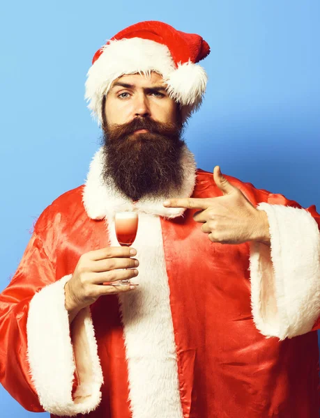 Красивий бородатий Санта-Клаус чоловік з довгою бородою на серйозному обличчі, тримає келих алкогольного пострілу в різдвяному або різдвяному светрі та новорічному капелюсі і показує на ньому на синьому студійному фоні — стокове фото