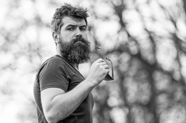 Jeune homme barbu musclé debout dans la vue de côté du parc, concept de mode de vie actif. Bel homme aux yeux bleus enlevant ses lunettes de soleil — Photo