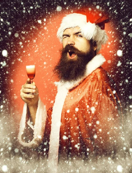 Guapo barbudo hombre de Santa Claus con barba larga en la cara divertida sosteniendo vaso de trago alcohólico en navidad o suéter de Navidad y sombrero de año nuevo en fondo rojo estudio —  Fotos de Stock