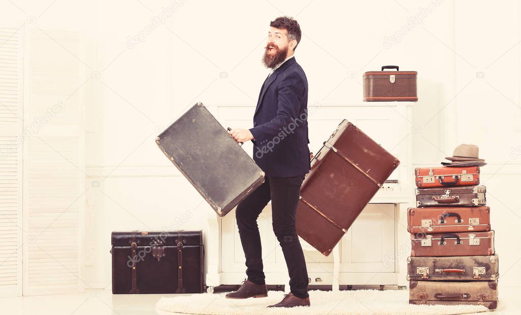 Man with beard and mustache wearing classic suit delivers luggage, luxury white interior background. Macho attractive, elegant on smiling face carries vintage suitcases. Butler and service concept