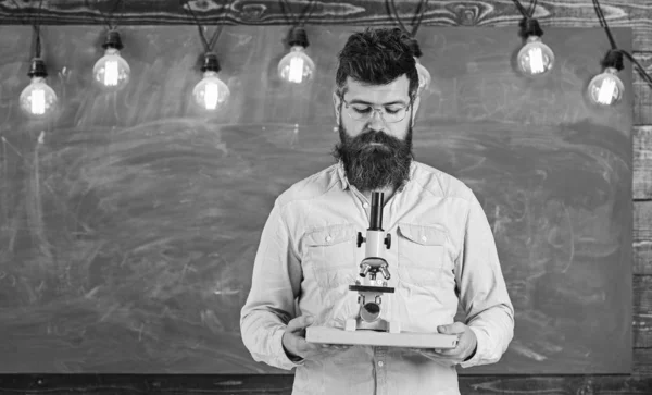 Man with beard and mustache on busy face. Teacher in eyeglasses holds book and microscope. Scientist holds book and microscope, chalkboard on background. Scientific research concept — Stock Photo, Image