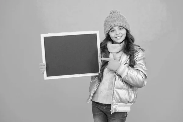 Concept de promotion. Enfant joyeuse promotion. Souriant fille porter tenue d'hiver montrer vide espace de copie tableau. Informer la communauté des enfants. Enfant avec tableau noir. Promotion des produits. Événement de promotion enfant — Photo