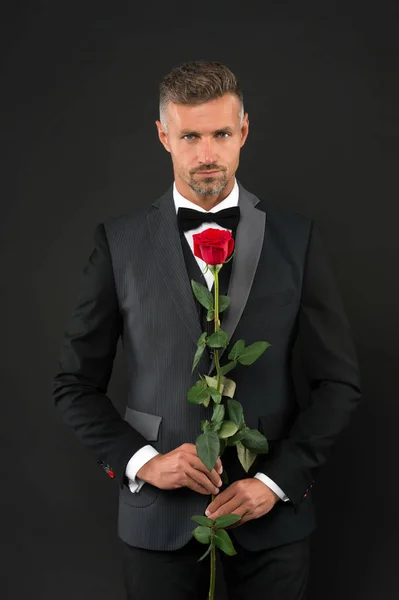 Ένας άντρας ντυμένος με σμόκιν και τριαντάφυλλο. Γαμπρός με γραβάτα και κόκκινο τριαντάφυλλο. Η κομψή Βαλεντίνα της. Ο άντρας με το σμόκιν κρατάει την Ρόουζ. Ο άντρας με το σμόκιν προσφέρει γάμο. πίσω παλτό ή frac. κόκκινο τριαντάφυλλο σύμβολο της αγάπης. ρομαντική αγάπη — Φωτογραφία Αρχείου