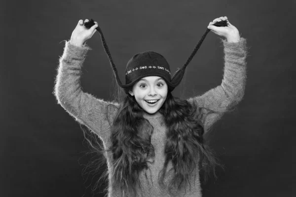 Zahřej zimu. Šťastné dítě v zimě. Dívka úsměv v pleteném klobouku na červeném pozadí. Malý módní model si užívá zimního stylu. Útulné zimní oblečení pro děti — Stock fotografie