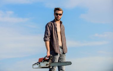 Elinde elektrikli testereyle oduncu. Tehlikeli bir iş. Kendine güveniyorsun. Riskli öfke. Güçlü elektrikli testere. Keskin bıçak. Testereli mavi gökyüzü arkaplanı olan yakışıklı bir adam. Bahçıvan oduncu ekipmanı.