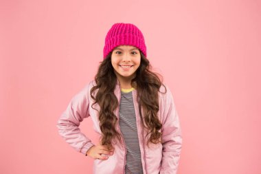 Sıcak kış kıyafetleri içinde neşeli bir çocuk. Çocuklar için mevsimlik moda. Küçük pembe duvar. Küçük kız kabarık ceket ve örgü şapka. Soğuk havada sağlık hizmeti. Havası nasıl olursa olsun