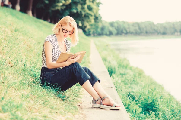 Een vrouw droomt van vakantie. Literatuur voor zomervakantie. Rust ontspannen en hobby. Zomervakantie. Meisje zit op groen gras in de buurt van de rivier en lees boek. Meisje ontspannen aan de rivier na werkdag — Stockfoto