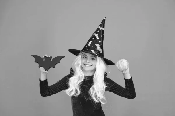 En riktig häxa i förklädnad. Glad liten häxa orange bakgrund. En liten häxa håller fladdermus och pumpa. Halloween barn leende i häxa hatt. Häxeri och magi. Vacker och mystisk — Stockfoto