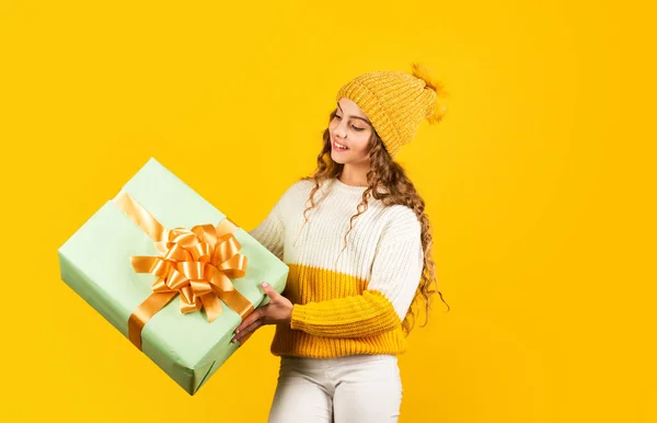 Она любит Рождество. Рождественская распродажа подарков. Счастливого Рождества вам. скидки на зимний отдых. Радостная маленькая девочка вязала шляпу и свитер. Ребенок держит коробку на желтом фоне. С Новым годом — стоковое фото
