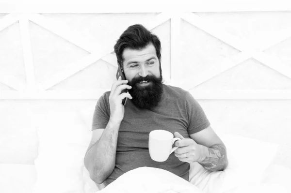 Hello dear. Bearded man using mobile technology in bed. Handsome guy talking on phone and drinking coffee at home. Modern life new technology. Technology concept. Pleasant conversation. Good morning