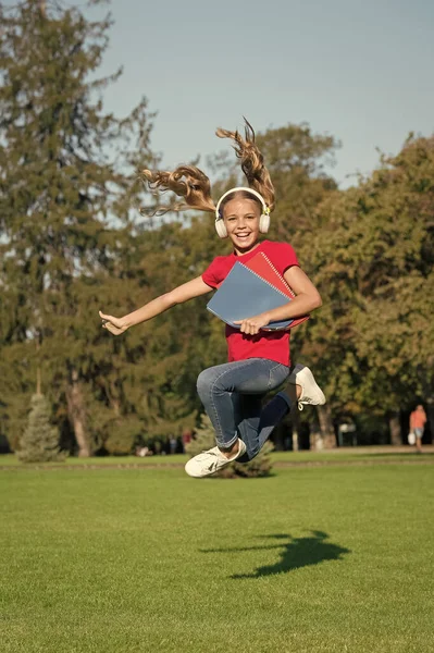 Moudrá volba pro fanatiky zvuku. Šťastné dítě skákat na zelené trávě. Malá holka poslouchá audioknihu ve sluchátkách. Tištěné a zvukové materiály pro vzdělávání. Zvukové příslušenství — Stock fotografie