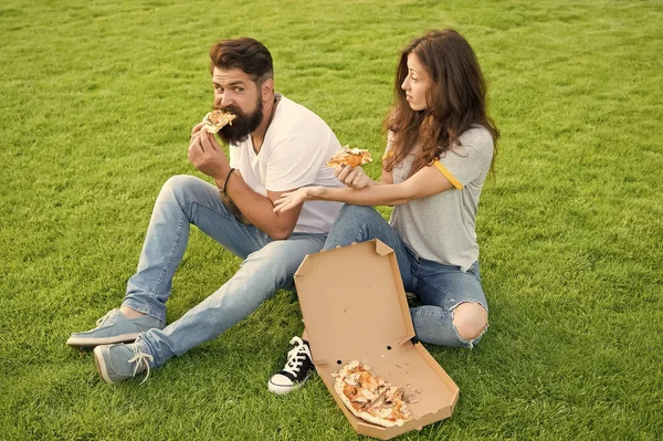 Hungry students sharing food. Pure enjoyment. Couple eating pizza relaxing on green lawn. Fast food delivery. Bearded man and woman enjoy cheesy pizza. Couple in love dating outdoors with pizza