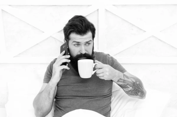 Bearded man using mobile technology in bed. Handsome guy talking on phone and drinking coffee at home. Modern life new technology. Technology concept. Pleasant conversation. Good morning. Hello dear
