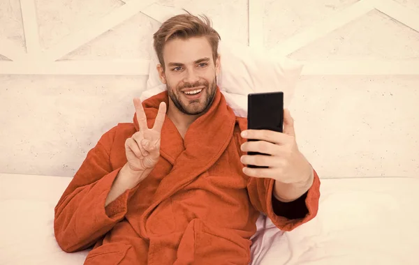自拍明星。快乐的男人在床上拿着智能手机自拍。英俊的男人对着手机里的自拍相机微笑着。在卧室享受自拍的乐趣 — 图库照片