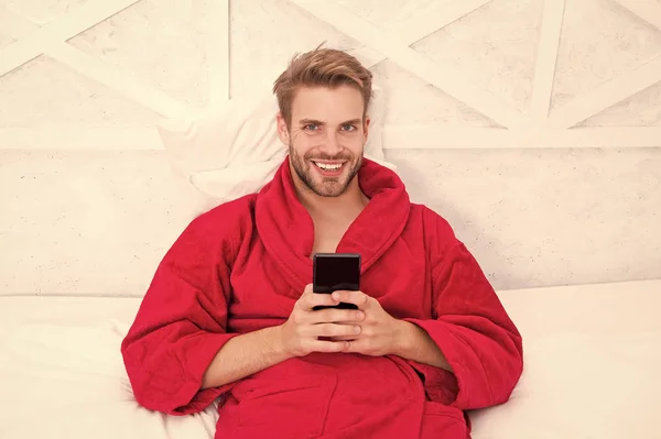 过着流动的生活。快乐的家伙在床上用移动设备。英俊的男人手里拿着手机笑着。家庭移动通信和无线技术 — 图库照片