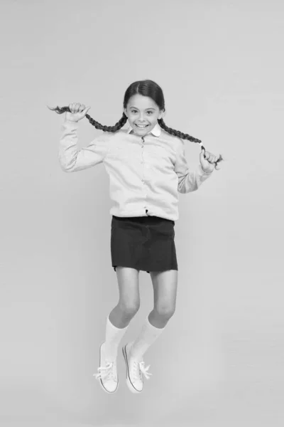 Ella es hiper. Feliz niño enérgico saltando sobre fondo amarillo. Niña con trenzas de pelo largo sintiéndose enérgica. Pequeño escolar que comienza enérgicamente el año escolar. Activo y enérgico —  Fotos de Stock