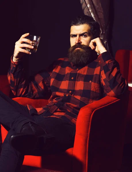Bearded man hipster holding glass of whiskey in red chair — Stock Photo, Image
