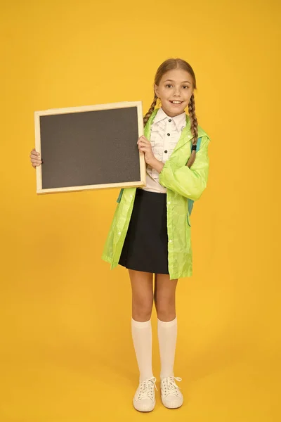 広告の配置。小さな子供は黄色の背景に関する情報のために空白の黒板を保持します。小さな女子高生はきちんとした情報ボードで笑顔。幸せな情報を共有する。あなたの情報、コピースペース — ストック写真