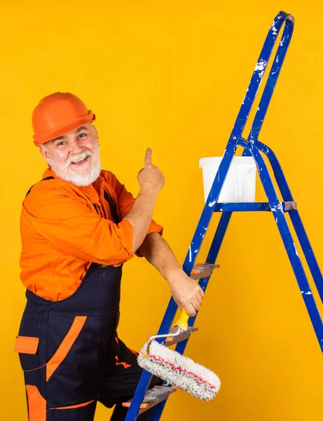 Це буде досконалим. старший художник використовує ролик на сходах. фарбування стіни жовтим. професійний художник у робочому одязі. працівник малює стіну в кімнаті. чоловічий декораторський живопис з роликом — стокове фото