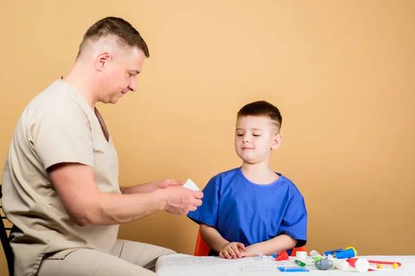 Eerste hulp. Medicijnconcept. Een kleine dokter zit aan tafel, medisch gereedschap. Gezondheidszorg. Medisch onderzoek. Jongen schattig kind en zijn vader arts. Ziekenhuismedewerker. Medische hulp. Medische verzekering — Stockfoto