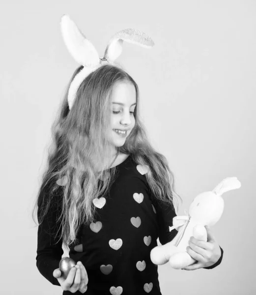 Пасхальные аксессуары для празднования в стиле. Очаровательная девушка с кроличьими ушами. Милая девушка в стиле Пасхи. Девочка играет с пасхальным яйцом и кроличьей игрушкой. Маленькая девочка с цветным яйцом и пасхальным кроликом — стоковое фото