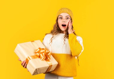 Küçük kız, sarı arka planda kurdelesi olan hediye kutusunu tutuyor. Kızım için Noel hediyesi. Sürprizlerin. Genç kız tatil hediyesi aldı. En iyi Noel hediyeleri. Çocuk hediyeyi açtığı için heyecanlı.