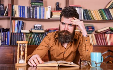 Toplu yüz okuma kitabı okuyan, ders çalışan, arka planda kitap rafları olan bir adam. Adam elinde kupa ve kum saatiyle masada oturuyor. Kütüphanede çalışan sakallı bir öğretmen ya da öğrenci. Kişisel eğitim kavramı.