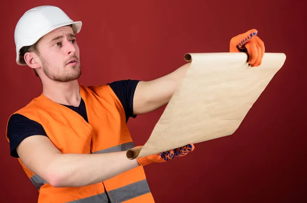 Engenheiro, arquiteto, construtor em face estrita mantém o velho modelo nas mãos, supervisiona o canteiro de obras. Homem de capacete, chapéu duro mantém plano de construção, controla obras, fundo vermelho. Conceito de Foreman . — Fotografia de Stock