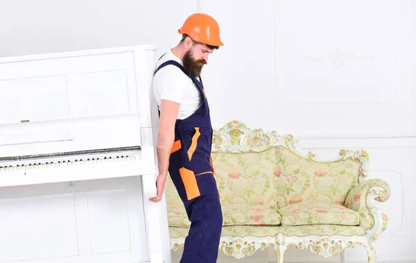 El cargador mueve el instrumento de piano. Concepto de cargas pesadas. El mensajero entrega muebles, se muda, se traslada. Hombre con barba trabajador en casco y overol se levanta, esfuerzos para mover el piano, fondo blanco . — Foto de Stock