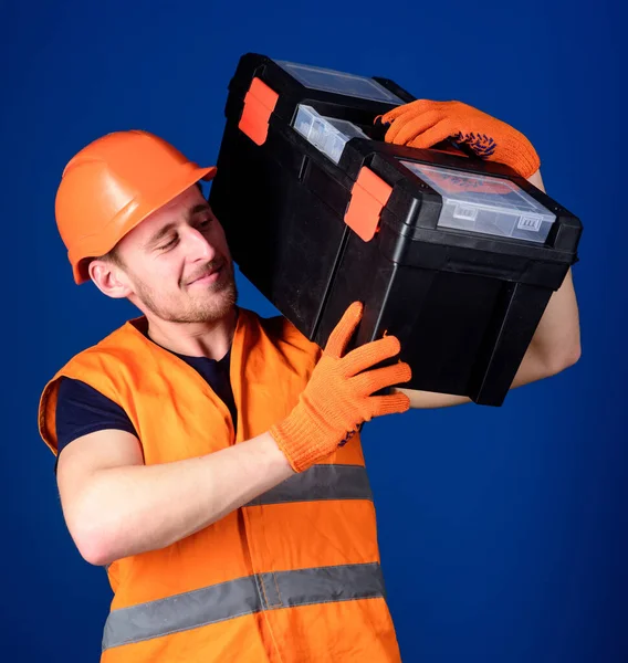 Trabalhador, reparador, reparador, construtor forte no rosto sorridente alegre carrega caixa de ferramentas no ombro, pronto para trabalhar. Conceito de serviço de reparação. Homem de capacete, chapéu duro segura caixa de ferramentas, fundo azul . — Fotografia de Stock