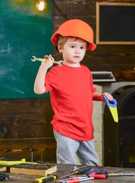 小木匠在车间帮忙.孩子在玩手锯和扳手。戴着橙色头盔的可爱男孩站在桌子后面，手里拿着工具 — 图库照片