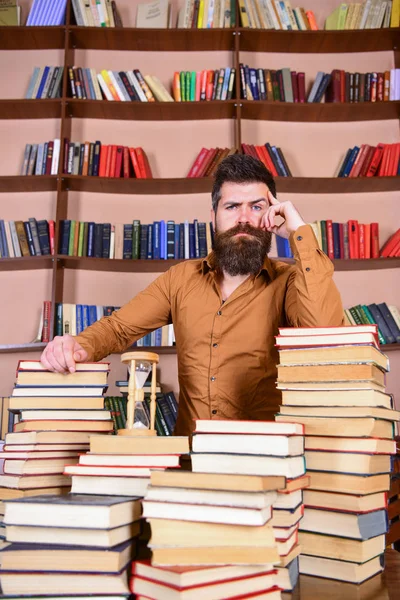 Учитель или ученик с бородой стоят за столом с книгами, обезглавленными. Библиотечная концепция. Человек на вдумчивом лице стоит между грудами книг, во время учебы в библиотеке, книжные полки на заднем плане . — стоковое фото