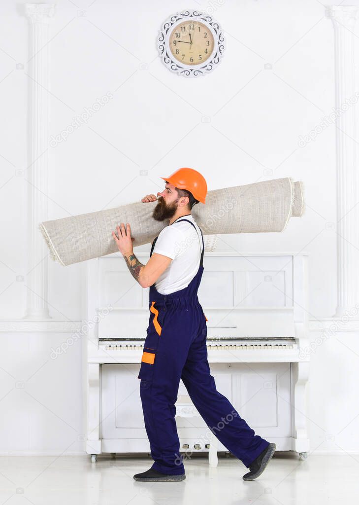 Relocating concept. Courier delivers furniture in case of move out, relocation. Loader wrapped carpet into roll. Man with beard, worker in overalls and helmet carries rolled carpet, white background.