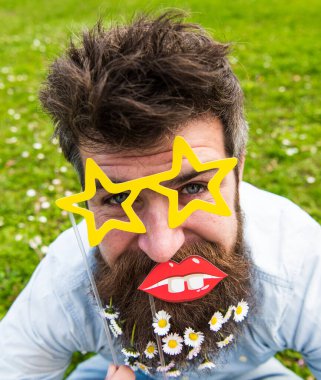 Neşeli yüzlü sakallı hippi yıldız şeklinde gözlük ve dudaklarla poz veriyor. Adam sakallı papatya çiçekleri ile güzel görünüyor. Sakallı ve bıyıklı bir adam, yeşil bir geçmişi var. Fotoğraf durgunluğu kavramı.
