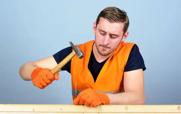 Handwerkliches Konzept. Tischler, Holzarbeiter auf konzentriertem Gesicht hämmert Nagel in Holzbrett. Mann, Handwerker in heller Weste und Schutzhandschuhen, hellblauer Hintergrund — Stockfoto