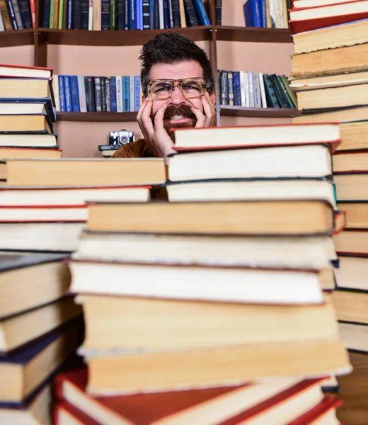 Homem no rosto nervoso entre pilhas de livros na biblioteca, estantes de livros no fundo. Conceito de descoberta científica. Professor ou estudante com barba usa óculos, senta-se à mesa com livros, desfocado . — Fotografia de Stock