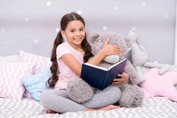 Όχι για μωρά. Κορίτσι παιδί κάθεται κρεβάτι διαβάστε το βιβλίο με αρκουδάκι. Ευχάριστη στιγμή στο άνετο υπνοδωμάτιο. Κορίτσι παιδί μακριά μαλλιά χαριτωμένο πιτζάμες χαλαρώσετε και να διαβάσετε το βιβλίο. Βιβλίο για την αγάπη. Αγαπημένο παραμύθι. Κλείσε τα μάτια σου. — Φωτογραφία Αρχείου