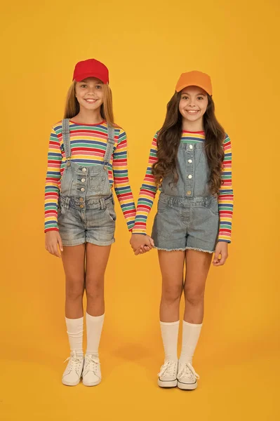 Les amis restent ensemble. Filles heureuses amis tenant la main sur fond jaune. Adorables petits amis appréciant l'amitié. Petits amis souriant en salopettes à la mode et casquettes élégantes — Photo