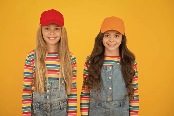 Hip hop girls. Little girls smiling on yellow background. Happy small girls wearing baseball caps in casual style. Adorable caucasian girls with long loose hair — Stock Photo, Image