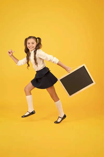 Pouco e enérgico. Criança carregando quadro preto no fundo amarelo. Pequena colegial com quadro preto para propaganda escolar. Menina com quadro vazio para o seu texto, espaço de cópia — Fotografia de Stock