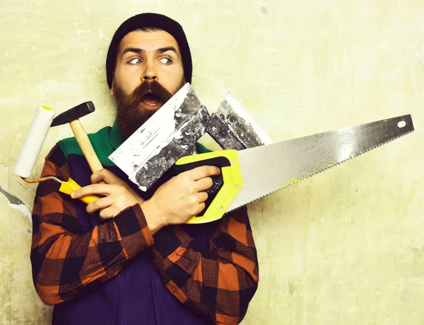 Бородатый человек держит различные строительные инструменты с удивленным лицом — стоковое фото