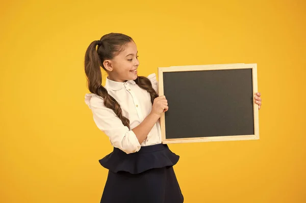 Menina pequena, grande sonho. Criança pequena segurando placa preta para propaganda escolar no fundo amarelo. Criança pequena com quadro-negro vazio para o seu texto. Pequena estudante segurando quadro negro, espaço de cópia — Fotografia de Stock