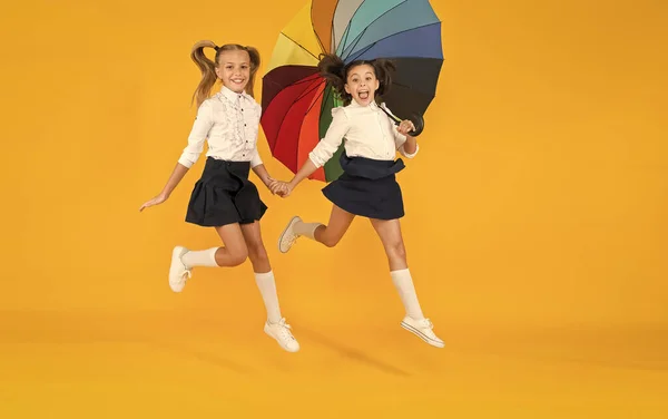 Back to school at any weather. Little girls of primary school age smiling under colorful umbrella on yellow background. Happy small children hurry to school. Enjoying school holidays in autumn