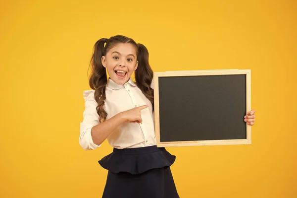 情報を見てください。黄色の背景に関する情報のために空白の黒板に指を指している小さな子供。学校の情報のためのきちんとした黒板を持つ小さな女の子。あなたのための情報、コピースペース — ストック写真
