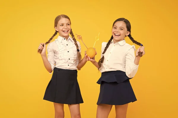 School dieting. Primary school pupils drinking juice from orange fruits on yellow background. Small children enjoying healthy school snack. Little girls with vitamin rich drink wearing school uniform — Stock Photo, Image
