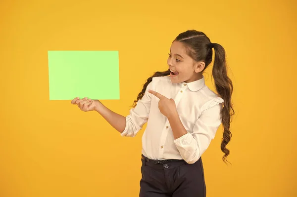 Gardez un œil sur l'idée. Fille heureuse pointant du doigt une certaine idée sur fond jaune. Petit enfant mignon souriant d'idée de génie. Petit enfant tenant du papier vide pour votre idée, copiez l'espace — Photo