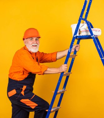 Boyacı merdiveni. Onarım yapmak için. İş elbiseli bir adam boş odaya duvar boyuyor. Boyalı kovayla şapkalı bir ressam. İnşaat işçisi boyama cephesi. termal yalıtım işe yarıyor