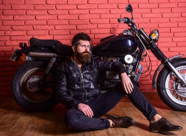 Sakallı bir adam, deri ceketli bir motorsikletli, garajda, tuğla duvarlı arka planda. Acımasız motorcu konsepti. Hipster, sert yüzlü, deri ceketli vahşi motorcu motosikletin yanına otur..