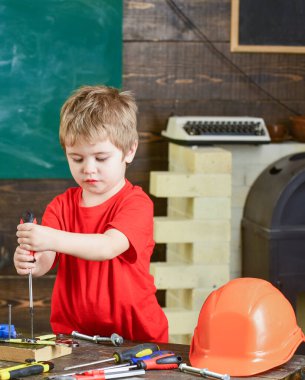 Odaklanmış çocuk iki eliyle tahtaya vida bağlıyor. Sarışın çocuk tamirat atölyesinde çalışıyor. Anaokulu öğrencisi yeni yetenekler kazanıyor