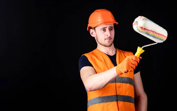 Decorator schilderen muur, kopieerruimte. Man in helm, harde hoed houdt verfroller, zwarte achtergrond. Werknemer, schilder, decorateur, aannemer, bouwer op serieus gezicht. Verf en renovatie concept. — Stockfoto